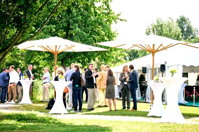 Verschiedene Personen stehen auf Rasen an Stehtischen unter Sonnenschirmen