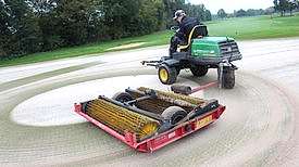 Person sitzt auf Aufsitzrasenmäher mit Anhänger und verstreut Sand auf Golfgrün