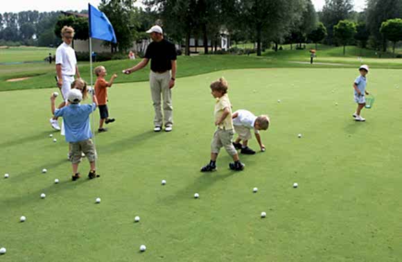 Mehrere Kinder stehen mit Golftrainer auf Putting Grün des Bambiniplatzes, Bälle liegen auf dem Boden