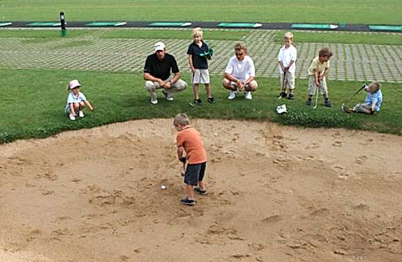 Kind steht im Bunker mit Golfball und Schläger während Kinder und Trainer außen herum stehen